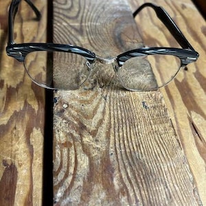 May include: Black and silver-rimmed eyeglasses with clear lenses. The glasses have a classic design with a half-rimmed frame and a silver bridge. The temples are black. The glasses are resting on a wooden surface.