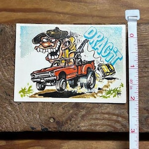 Puede incluir: Pegatina vintage con una ilustración de dibujos animados de una grúa roja con un conductor y la palabra "DRAG IT" en azul. El conductor lleva un sombrero y un coche amarillo está siendo remolcado.