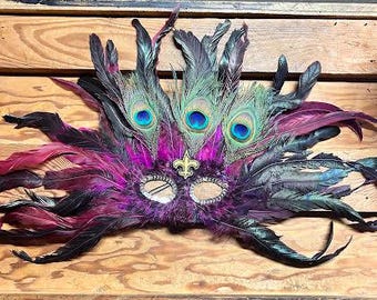 May include: A decorative Mardi Gras mask with peacock feathers and a fleur-de-lis emblem. The mask features a purple base with black and magenta feathers, and three peacock feathers with blue and green accents.