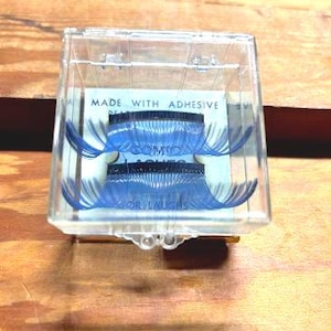 Könnte beinhalten: Ein durchsichtiger Plastikbehälter mit einem blauen und weißen falschen Wimpern-Set. Der Behälter trägt ein Etikett mit der Aufschrift "Made with Adhesive Real Lashes for Lashes".