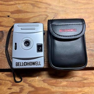 May include: A silver Bell+Howell camera with a black strap next to a black leather case. The camera has a lens, a flash, and the brand name printed on the front. The case has the brand name in red.