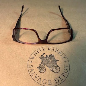 May include: Tortoiseshell-patterned rectangular spectacles with thin frames. Below the glasses is a circular stamp featuring a rabbit pulling a carriage and the text "WHITE RABBIT SALVAGE DEPOT" printed in a dark ink on a light brown surface.