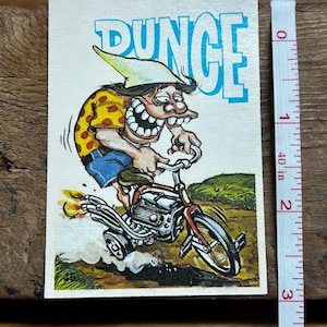 Puede incluir: Postal vintage con un personaje de dibujos animados montado en una motocicleta. El personaje tiene una gran sonrisa, viste una camisa amarilla con lunares rojos, pantalones cortos azules y un gorro de burro. La palabra "DUNCE" está en letras azules grandes.