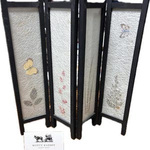 May include: A four-panel folding screen with a black frame and white panels. The panels feature decorative elements like butterflies, leaves, and floral designs. The screen is freestanding and has a small white card with text at the base.