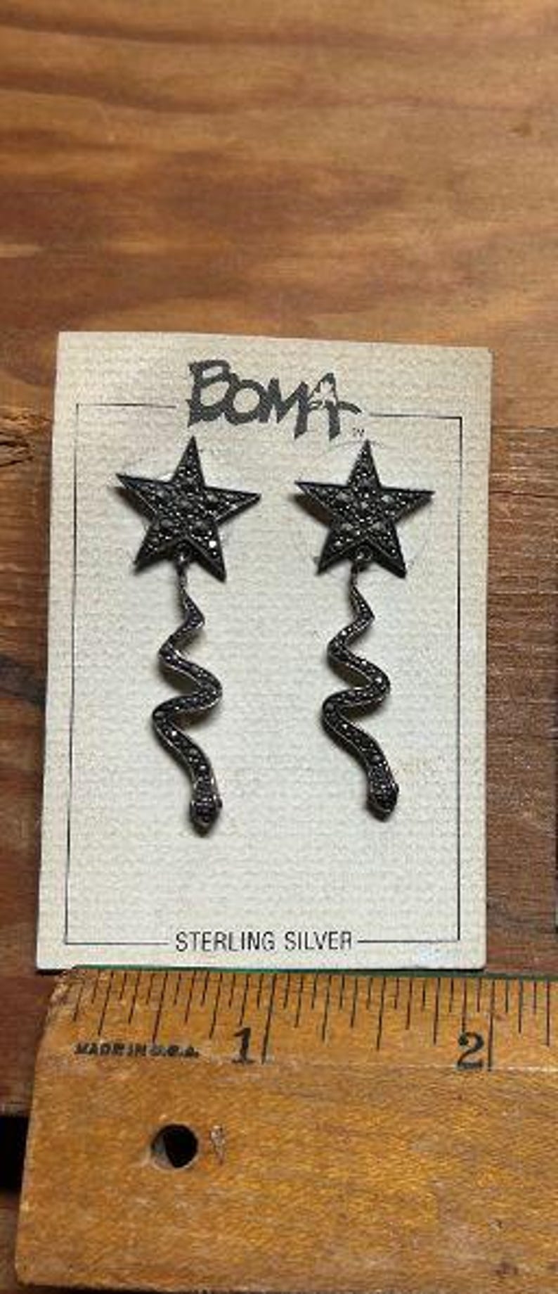 May include: A pair of sterling silver earrings with a star design and a dangling, twisted, chain-like detail. The earrings are on a white card with the word "BOMA" printed on it.