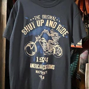 May include: Dark navy blue t-shirt featuring a motorcycle graphic with the text "THE ORIGINAL SHUT UP AND RIDE" and "1974 AMERICAN CUSTOMS". The design is in white and light blue.