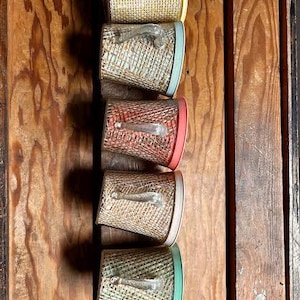 Puede incluir: Una colección de cinco recipientes vintage tejidos con asas transparentes y bordes de colores. Los recipientes están dispuestos verticalmente sobre un fondo de madera. Los bordes son amarillos, azules, rojos, beiges y verdes.