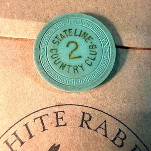 May include: A round, light blue poker chip featuring a Greek key border pattern. The centre displays the text STATELINE COUNTRY CLUB in a circular arrangement with the number 2 printed in the middle. The chip has a textured, matte finish.