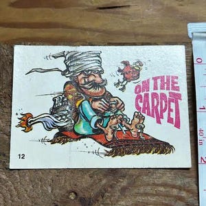 Puede incluir: Una tarjeta vintage con una ilustración de dibujos animados de un hombre con turbante, sentado en una alfombra mágica. El texto "ON THE CARPET" está en rosa. La tarjeta tiene el número 12 en la esquina inferior izquierda.