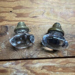 May include: Two clear, faceted glass door knobs with metallic brass-toned bases. Each knob features a multi-sided, geometric shape that reflects light, attached to a circular, stepped brass-coloured metal mounting piece at the top.