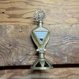 May include: An antique brass perfume bottle with a decorative floral top and a triangular mirror. The bottle has a flared base with cut-out designs and is set against a wooden background. The bottle is approximately 15 cm tall.
