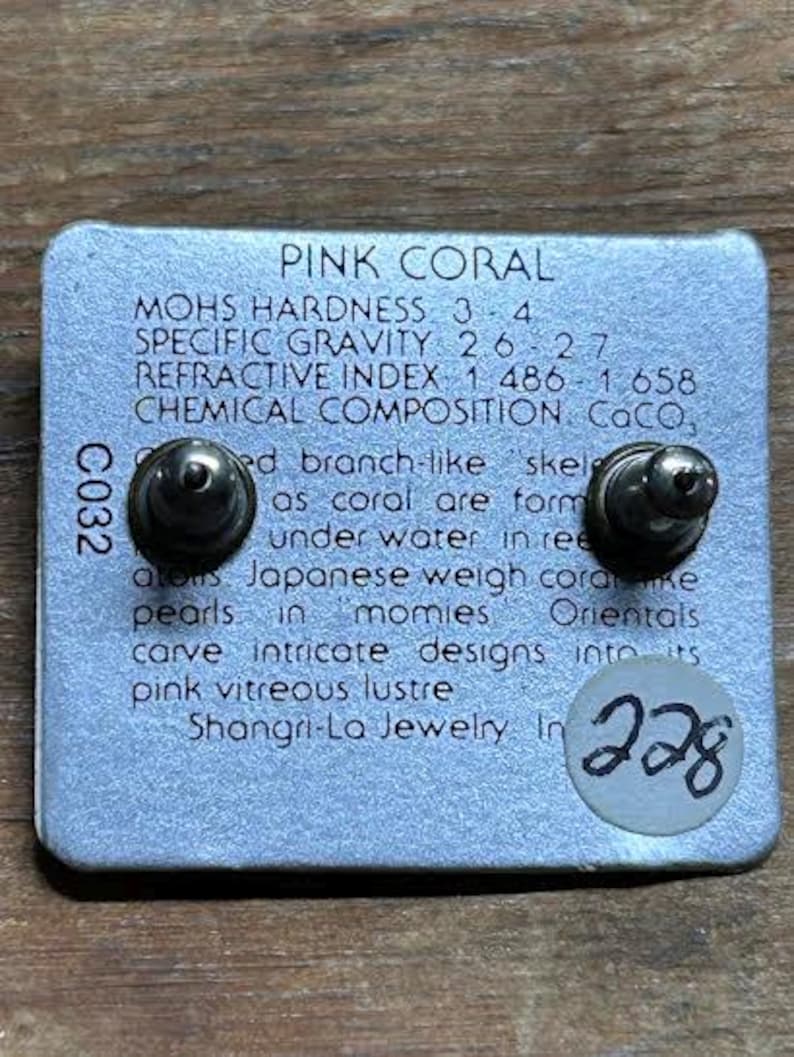 K&ouml;nnte beinhalten: Eine silberne Ohrringkarte mit zwei Ohrsteckern. Die Karte tr&auml;gt den Text "PINK CORAL" und Details &uuml;ber die Eigenschaften der Koralle. Ein kleiner Aufkleber mit der Nummer "228" befindet sich auf der Karte.