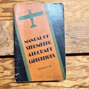 Puede incluir: Un libro negro y naranja titulado "Manual de carburadores de avión Stromberg" con una ilustración de avión verde en la portada. El libro está etiquetado como "Edición IV" y tiene una inscripción manuscrita.