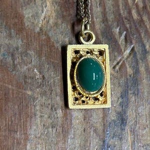 May include: A gold-colored pendant necklace with a rectangular frame and a green oval stone. The pendant hangs from a delicate chain. The frame has an openwork design.