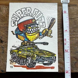 Puede incluir: Pegatina vintage de Super Fuzz que presenta un personaje de dibujos animados con un casco de policía, sosteniendo un bate, encima de un vehículo de estilo militar. El vehículo es verde oliva con la palabra «POLICE». La pegatina mide unos 5 cm de alto.