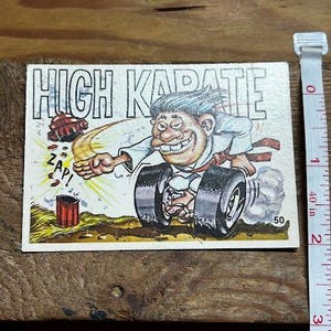 Puede incluir: Una tarjeta comercial vintage con la frase "HIGH KARATE". La caricatura representa a un hombre con uniforme de karate sobre ruedas, rompiendo un coche con un golpe de karate. La palabra "ZAP!" también es visible.