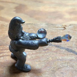 May include: A pewter figurine of a gnome holding a crystal ball and a wand. The gnome is wearing a pointed hat and a long coat. The figurine is standing on a wooden surface.