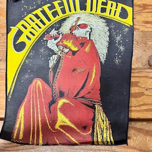 May include: A black patch featuring the words "Grateful Dead" in yellow, arched over an illustration of a skeleton figure in a red robe. The skeleton has a white afro and is holding a violin. The background is black with yellow and white accents.