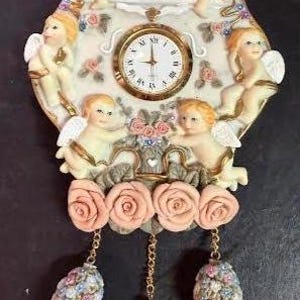 May include: A decorative wall clock with a cream-coloured base, featuring cherubic figures and floral accents. The clock face is gold-toned with Roman numerals. The clock has three decorative hanging elements.