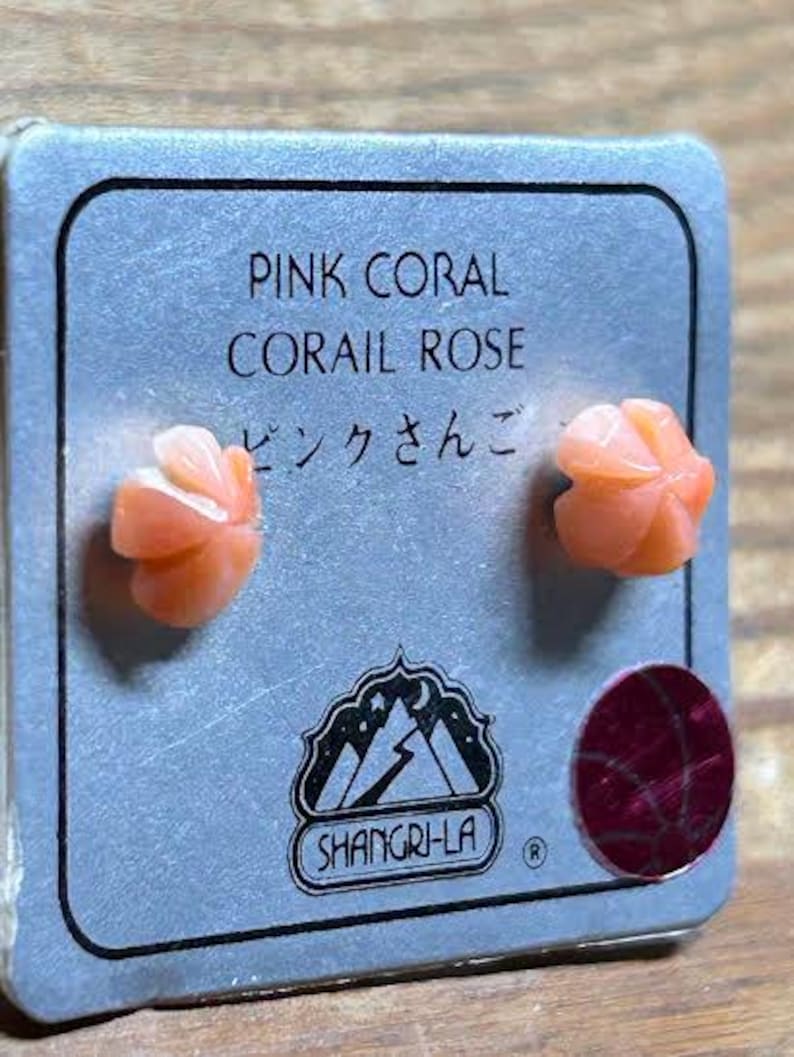 K&ouml;nnte beinhalten: Ein Paar rosa Korallenrosen-Ohrstecker auf einer grauen Karte. Auf der Karte steht "PINK CORAL CORAIL ROSE" und japanische Schriftzeichen. Die Ohrringe sind hellorange und rosenf&ouml;rmig.