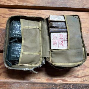 Puede incluir: Botiquín de primeros auxilios de lona verde oliva abierto con cremallera. El kit contiene vendas, apósitos y suministros de primeros auxilios. El kit está abierto, revelando el contenido, sobre un fondo de madera.