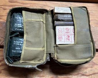 Puede incluir: Botiquín de primeros auxilios de lona verde oliva abierto con cremallera. El kit contiene vendas, apósitos y suministros de primeros auxilios. El kit está abierto, revelando el contenido, sobre un fondo de madera.