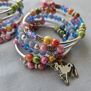 Peut inclure: Bracelets à plusieurs brins avec des perles colorées dans les tons de rose, bleu, vert, orange et jaune. Chaque bracelet a une bande argentée et un charm en forme de licorne blanche avec une crinière et une queue arc-en-ciel.