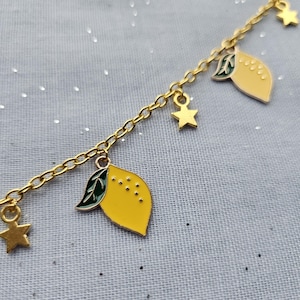 May include: Gold-coloured chain bracelet featuring yellow lemon charms and star-shaped charms. The lemons have green leaves and small black dots. The bracelet is on a light grey background.