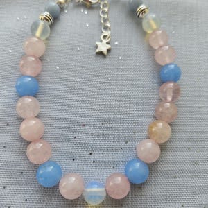 Può includere: Un braccialetto realizzato con perline di pietre preziose rosa, blu e trasparenti. Il braccialetto ha una chiusura argentata e un ciondolo a forma di stella.