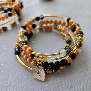 Puede incluir: Un conjunto de cuatro pulseras de brazalete doradas con cuentas negras, blancas y naranjas. Las pulseras tienen un dije en forma de corazón.