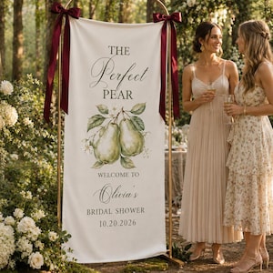 May include: A vertical white fabric banner on a gold metal stand, featuring a watercolor pear illustration and the text THE PERFECT PEAR, WELCOME TO Olivia's BRIDAL SHOWER, 10.20.2026. The banner is accented with dark red satin ribbons.