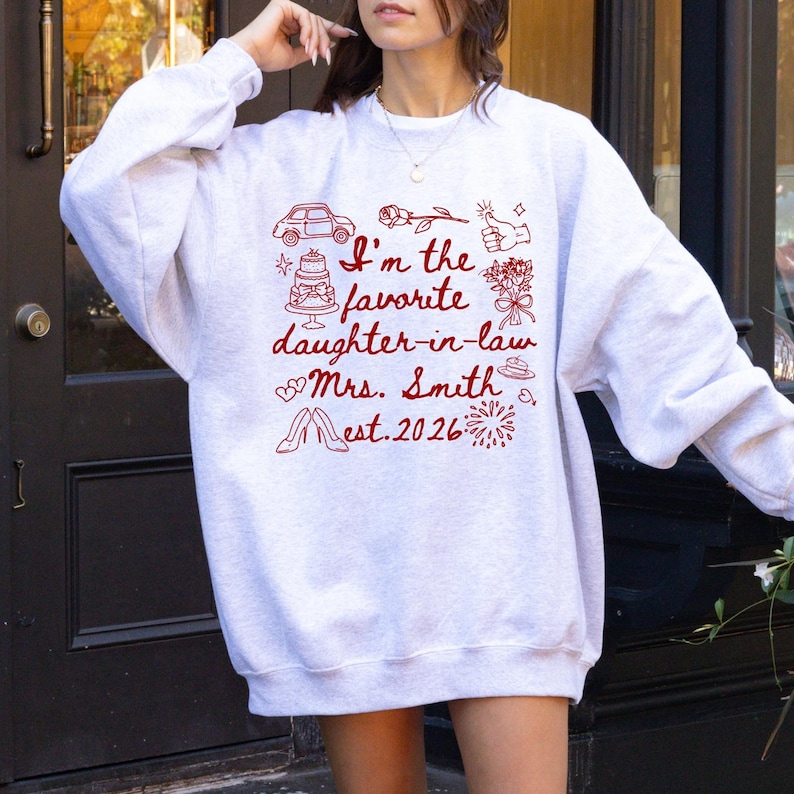 Op de afbeelding: Een lichtgrijze sweatshirt met een rode afbeelding met de tekst "I'm the favorite daughter-in-law Mrs. Smith Est. 2026" met illustraties van een auto, taart, bloemen en andere feestelijke iconen. De sweatshirt heeft een casual stijl.