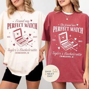 Könnte beinhalten: Zwei T-Shirts, eines in Elfenbein und eines in Karmesinrot, zeigen den Text "Found my PERFECT MATCH" und "She found her PERFECT MATCH". Beide Shirts haben eine Grafik einer Streichholzschachtel mit Herzen und dem Text "Taylor's Bachelorette, Charleston, SC, EST. 2023".
