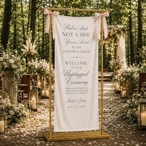Könnte beinhalten: Ein weißes Hochzeitsschild aus Stoff, das an einem goldfarbenen Metallständer mit rosa Schleifen hängt. Der Text lautet: Pick a Seat NOT A SIDE You are Loved BY THE GROOM & BRIDE WELCOME TO OUR Unplugged Ceremony, gefolgt von Anweisungen und den Namen Hailey & Justin.