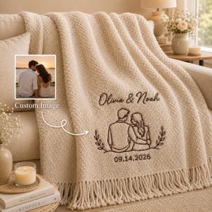 May include: A cream-coloured knitted throw blanket featuring a dark brown embroidered line art illustration of a couple sitting together. The text "Olivia & Noah" is embroidered above the illustration, with the date "09.14.2026" below it.