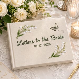 Puede incluir: Un libro cubierto de tela color crema con el título Letters to the Bride y la fecha 10.12.2026 bordados en hilo verde oscuro. La portada presenta ramas de lirio de los valles bordadas y una mariposa con contornos negros.