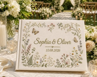 Könnte beinhalten: Ein rechteckiges Gästebuch mit einem cremefarbenen Stoffeinband, verziert mit gestickten Wildblumen, Grünpflanzen und zwei Schmetterlingen. In der Mitte steht der Text Sophia & Oliver sowie das Datum 11.08.2026 in grüner Schrift.