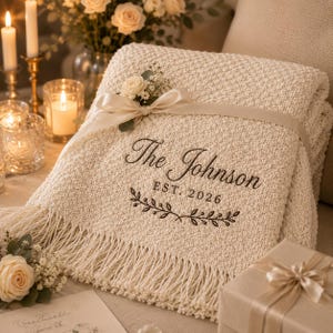 Könnte beinhalten: Eine cremefarbene, texturierte Strickdecke mit Fransenrand und einem darum gebundenen Satinband. Die Decke ist mit dem Text The Johnson EST. 2026 über einem dekorativen Zweigmotiv bestickt und mit einem kleinen Blumenarrangement verziert.