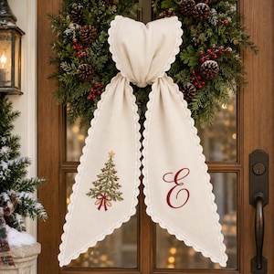 Peut inclure: Un accessoire de porte en tissu crème avec une bordure en dentelle festonnée et deux longs pans. Un pan présente un sapin de Noël brodé en vert avec une étoile dorée, tandis que l'autre porte une grande lettre E brodée en rouge.