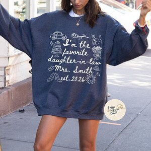 Op de afbeelding: Een donkergrijs sweatshirt met witte tekst en afbeeldingen. De tekst luidt "I'm the favorite daughter-in-law Mrs. Smith Est. 2026" met illustraties van een auto, taart, bloemen en schoenen. Het sweatshirt heeft een casual stijl.