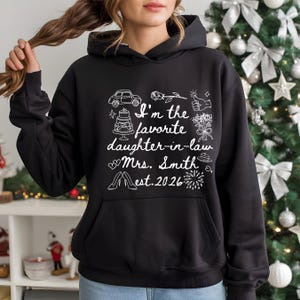 Op de afbeelding: Zwarte hoodie met witte tekst en illustraties. De tekst luidt "I'm the favorite daughter-in-law Mrs. Smith est. 2026" met tekeningen van een auto, taart, bloemen en andere iconen.