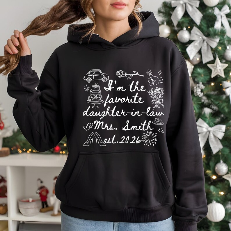 Op de afbeelding: Zwarte hoodie met witte tekst en illustraties. De tekst luidt "I'm the favorite daughter-in-law Mrs. Smith est. 2026" met tekeningen van een auto, taart, bloemen en andere iconen.