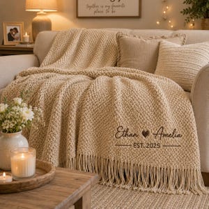 Op de afbeelding: Een beige gebreide plaid met franjes, voorzien van de geborduurde tekst "Ethan ♥ Amelia" en "EST. 2025" in donkerbruin cursief schrift. De deken heeft een textuur met een korrelig patroon en ligt gedrapeerd over een bank met bijpassende kussens.
