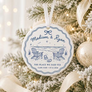 May include: A round white ceramic ornament with a blue scalloped border and a white ribbon hanger. The design features two beach chairs, a ring box, and an oyster shell on sand. Text reads: Madison & Ryan, THE PLACE WE SAID YES, CAPE COD 07.11.2026.