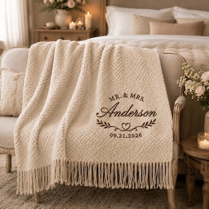 May include: A cream-coloured textured throw blanket with fringe trim, featuring brown embroidered text that reads MR. & MRS. Anderson above a heart and leaf design, with the date 09.21.2026 embroidered in brown thread at the bottom.