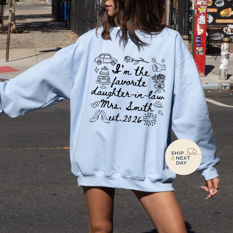 Op de afbeelding: Lichtblauw sweatshirt met de tekst "I'm the favorite daughter-in-law Mrs. Smith est. 2026" en diverse doodle-illustraties. Het ontwerp bevat een auto, taart, bloemen en andere feestelijke iconen.