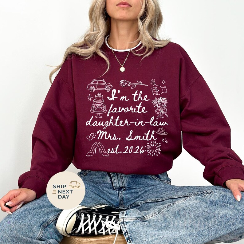 Op de afbeelding: Bordeaux sweatshirt met witte grafische tekst en illustraties. De tekst luidt "I'm the favorite daughter-in-law Mrs. Smith est. 2026" met afbeeldingen van een auto, taart, bloemen en schoenen. De sweatshirt is een ronde hals stijl.