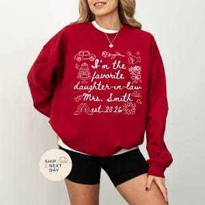 Op de afbeelding: Een rode sweatshirt met witte tekst en illustraties. De tekst luidt "I'm the favorite daughter-in-law Mrs. Smith est. 2026" met afbeeldingen van een auto, taart, bloemen en schoenen. De sweatshirt heeft een ronde hals.