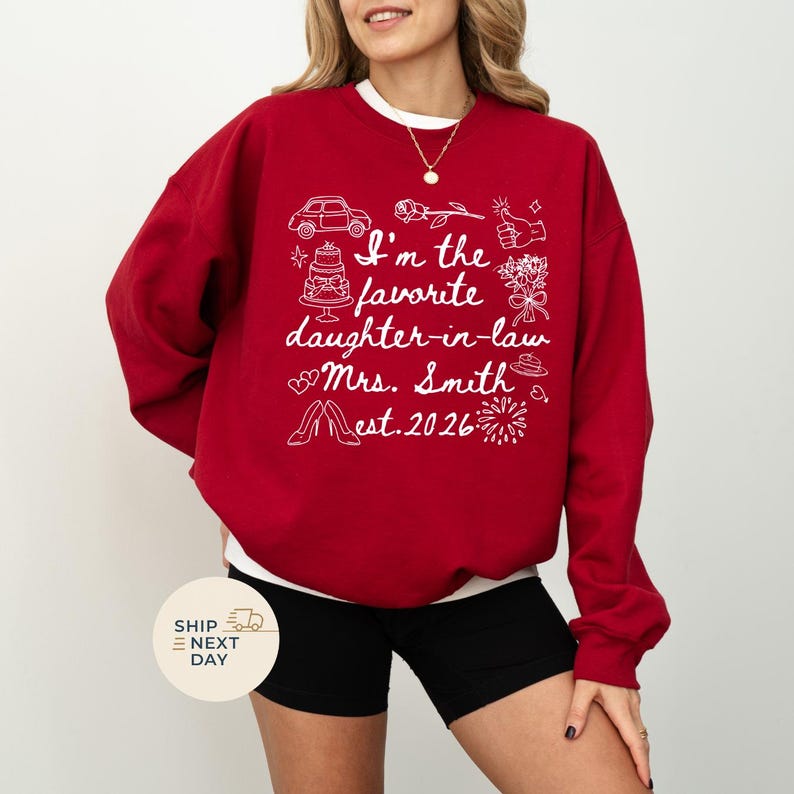 Op de afbeelding: Een rode sweatshirt met witte tekst en illustraties. De tekst luidt "I'm the favorite daughter-in-law Mrs. Smith est. 2026" met afbeeldingen van een auto, taart, bloemen en schoenen. De sweatshirt heeft een ronde hals.