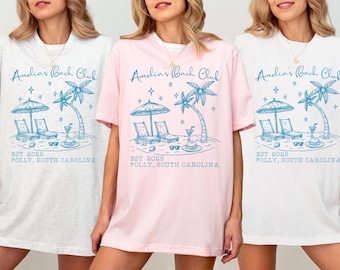 Może przedstawiać: Trzy koszulki z grafiką o tematyce plażowej. Projekt zawiera parasol, leżaki, palmy i tekst "Amelia's Bach Club EST 2025 FOLLY, SOUTH CAROLINA." Koszulki są w kolorze białym, jasnoróżowym i szarym.